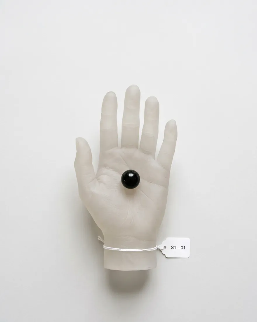 A frosted silicone hand, palm up, holding a black glass sphere. It slowly closes into a fist.